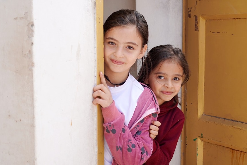 Two young girls smile at the camera from a doorway
