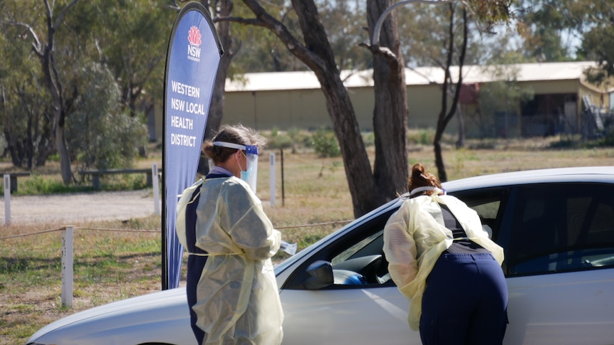 Two health workers in full PPE, one bending over to speak to the occupants of a car, the other standing by