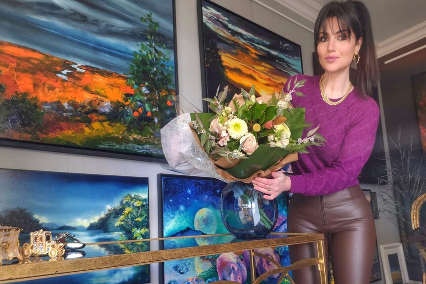 A woman with brown high ponytail and fuchsia jumper holds a bouquet of flowers and poses in front of her landscape paintings