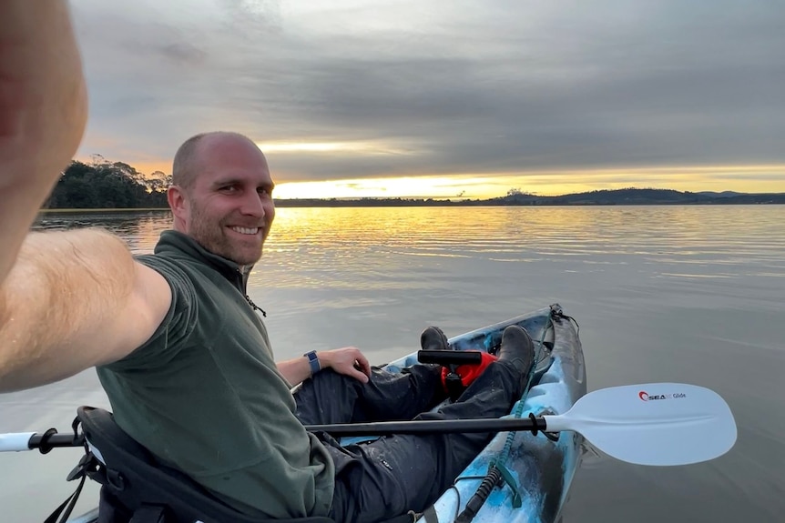 Man sitting in water craft smiling, he holds camera extended. In background is the ocean and a sunset or sunrise.