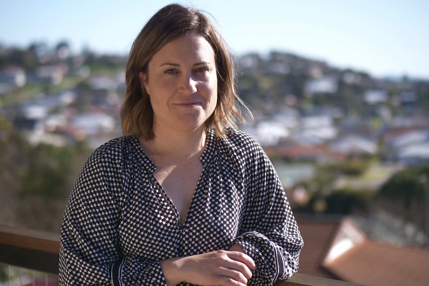 Young woman wearing black and white shirt standing on a balcony in the morning sun with a hill in the background.