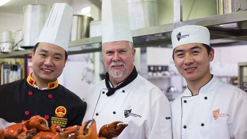 Three chefs stand in a row in a kitchen, holding a tray of orange crabs