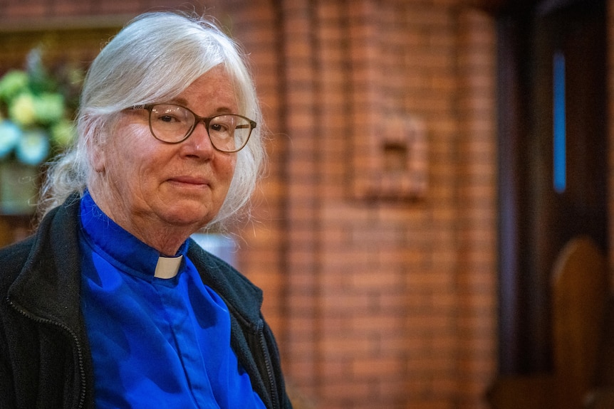 Reverend Fran Grimes, wearing glasses and a blue shirt with white collar, stands in a brick church.