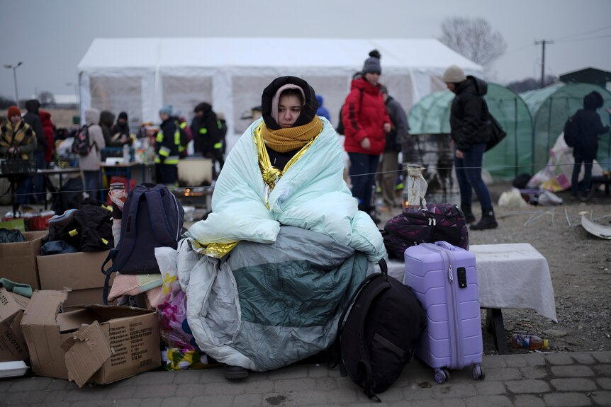 A woman sitting on a bench is wrapped in sleeping bags and blankets, in the background is a camp for people fleeing to Poland.