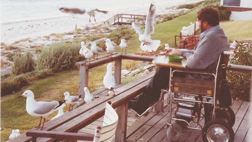 A man in a wheelchair on a balcony surrounded by seagulls and looking out at the ocean.