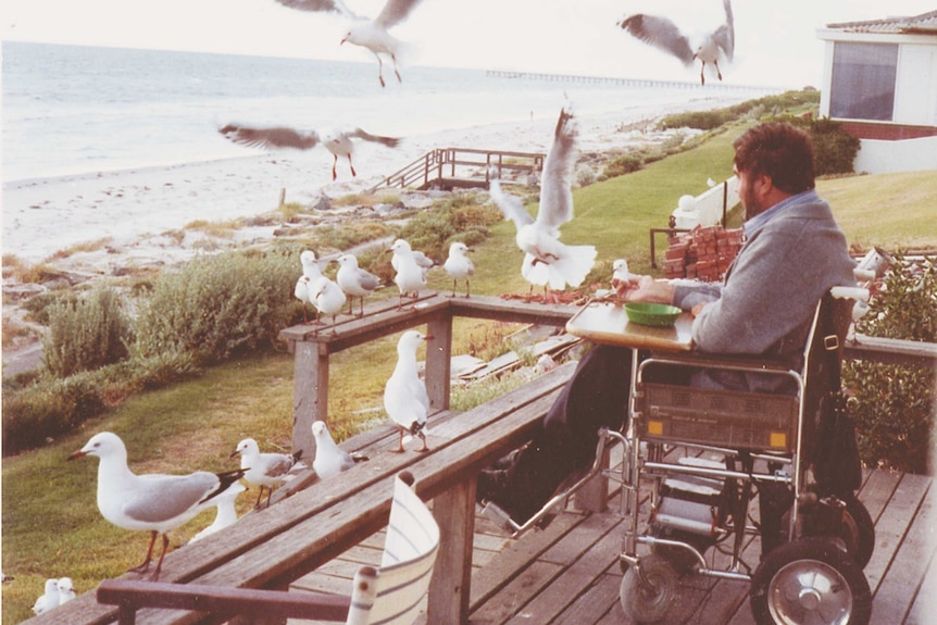 A man in a wheelchair on a balcony surrounded by seagulls and looking out at the ocean.