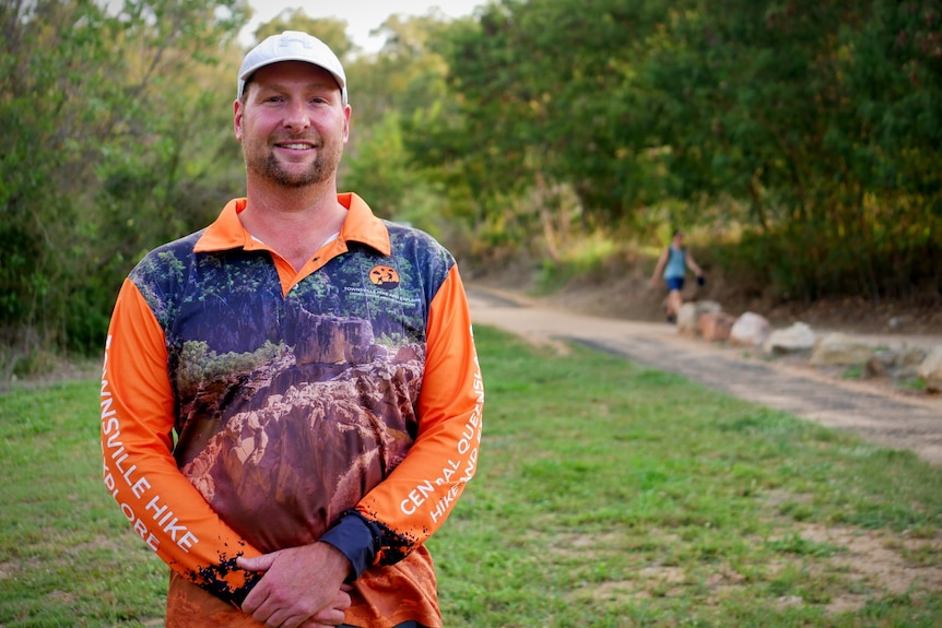 A man in an orange and black hiking shirt stands in front of a bush walking trail.