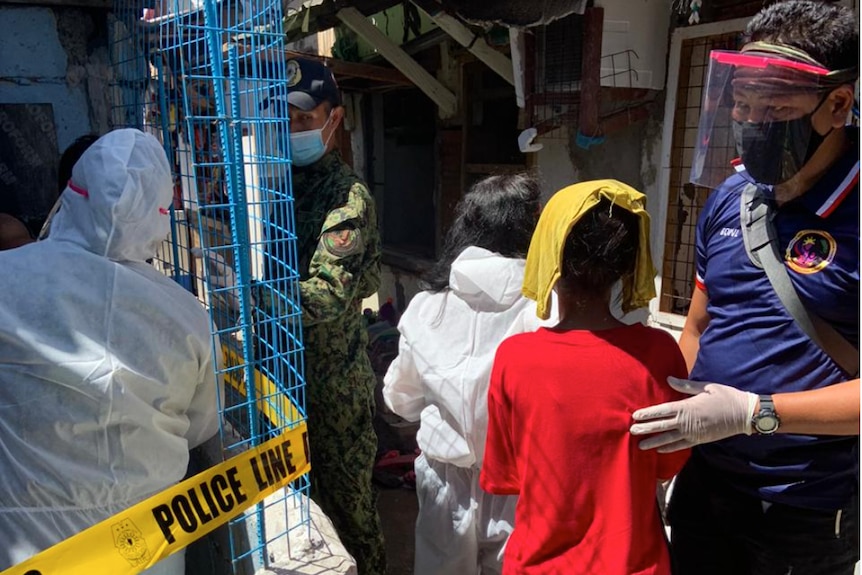 A man in PPE in a slum, with a young boy in front of him with his face covered by a towel