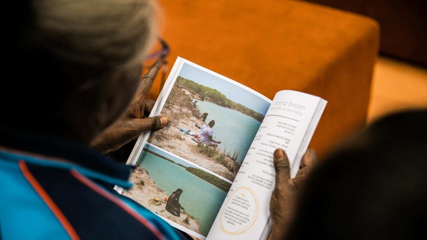 An aged Aboriginal woman looks at a page of the cookbook, with photos of young local children playing.
