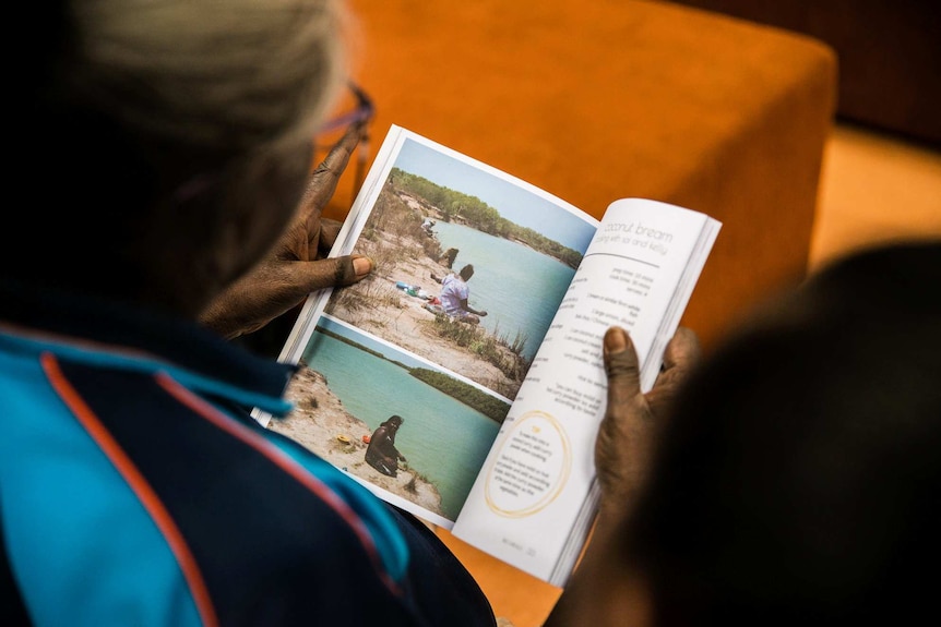 An aged Aboriginal woman looks at a page of the cookbook, with photos of young local children playing.