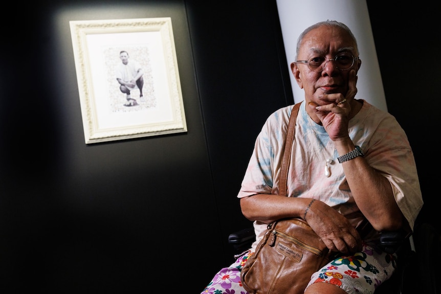 A man sitting in front of a black-and-white portrait handing on a wall in a gallery. He wears a pearl necklace.