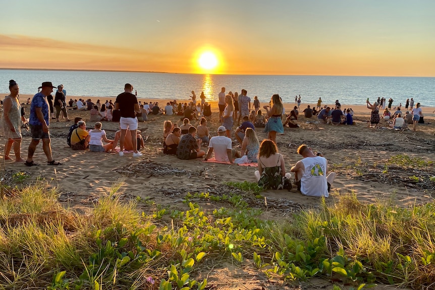 People sit on Mindil Beach as the sun sets over the sea. 