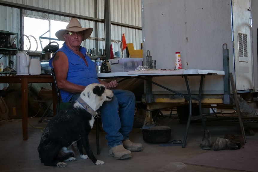 A man sits with his black and white dog, in a makeshift kitchen in a shed. 