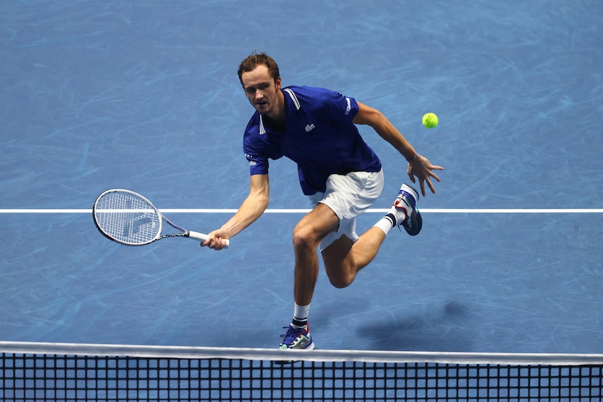 A Russian tennis player leans in toward the net as he tries to hit a crosscourt forehand during a match.