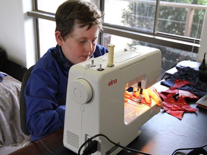 A boy sitting at a sewing machine in his bedroom, sewing a small hat.