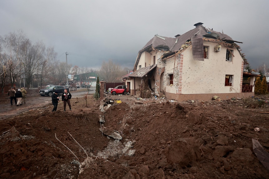 People stand by a very large hole in the ground next to a house that has suffered severe blast damage.