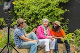 Three people sitting in garden laughing with lighting stand in shot.