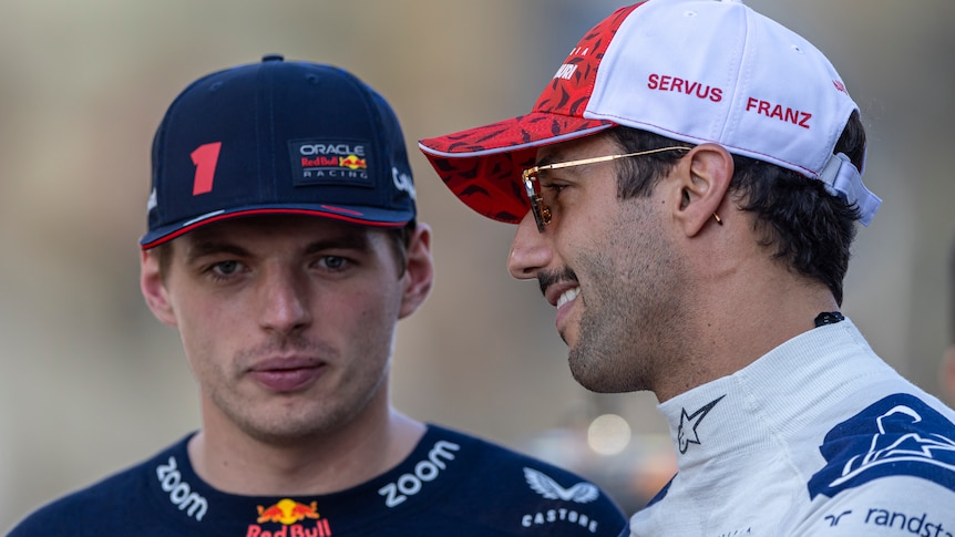 Two F1 drivers, one in blue and one in white, wearing caps, talking to each other before a race.
