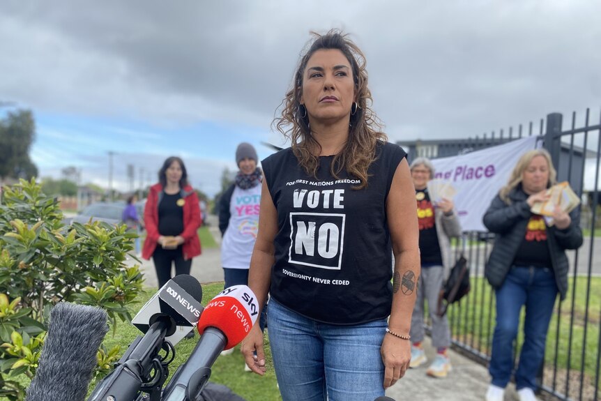 Lidia Thorpe stands in front of media microphones wearing a t-shirt that says 'vote no'
