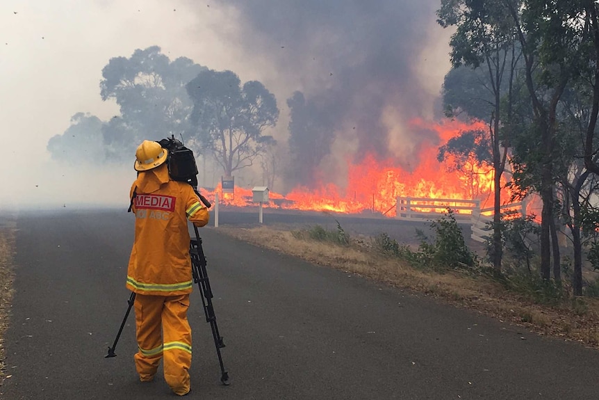 Cameraman in yellow protective suit and helmet filming flames and black smoke.