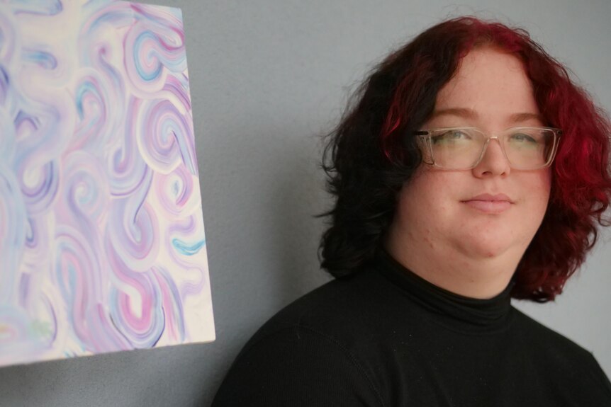 A teen with glasses and red and black hair poses in front of a pastel artwork