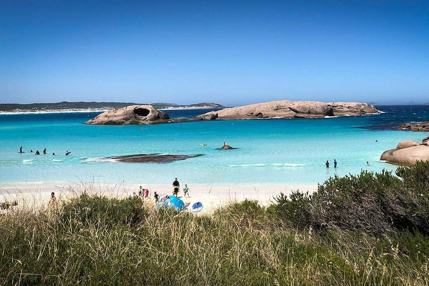 Bright blue waters and white sands of a beach in south-west WA with people on beach and in water and rocks in water