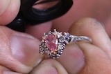 A close-up shot of a man's hands holding a pink diamond ring and a jeweller's magnifying glass with his face in the background.