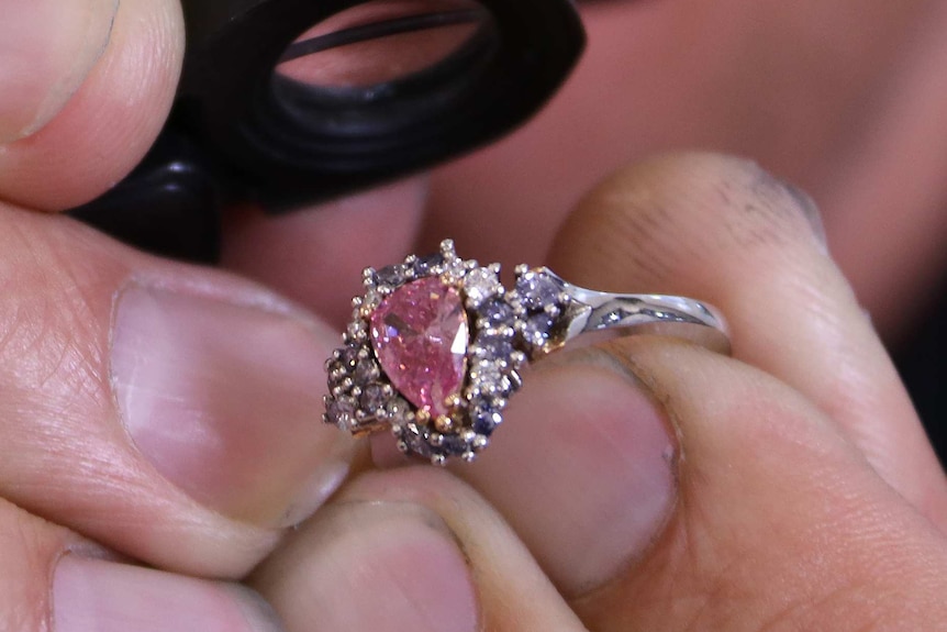 A close-up shot of a man's hands holding a pink diamond ring and a jeweller's magnifying glass with his face in the background.
