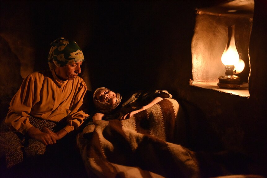 Inside a dark room a sitting woman with floral headscarf looks down at elderly woman laying down, both illuminated by oil lamp.