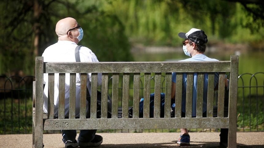 Two men wearing facemasks sit at the far ends of a park bench with greenery in the background.