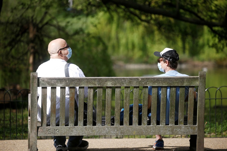 Two men wearing facemasks sit at the far ends of a park bench with greenery in the background.