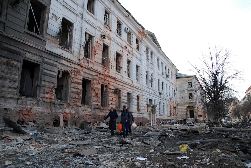 Two people walk through rubble next to a large building whose windows have blown and walls are peppered with holes.
