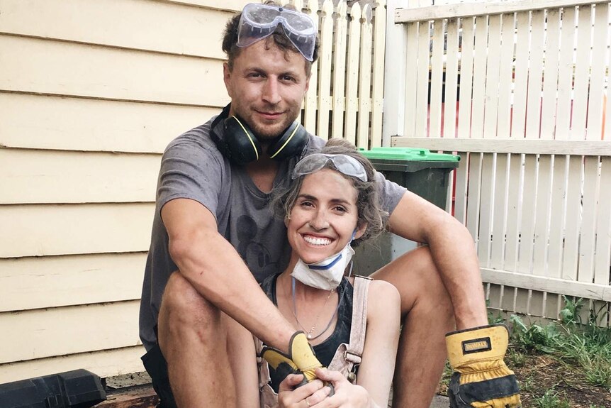 A young man and woman sitting outside a house smiling while covered in dirt from renovating