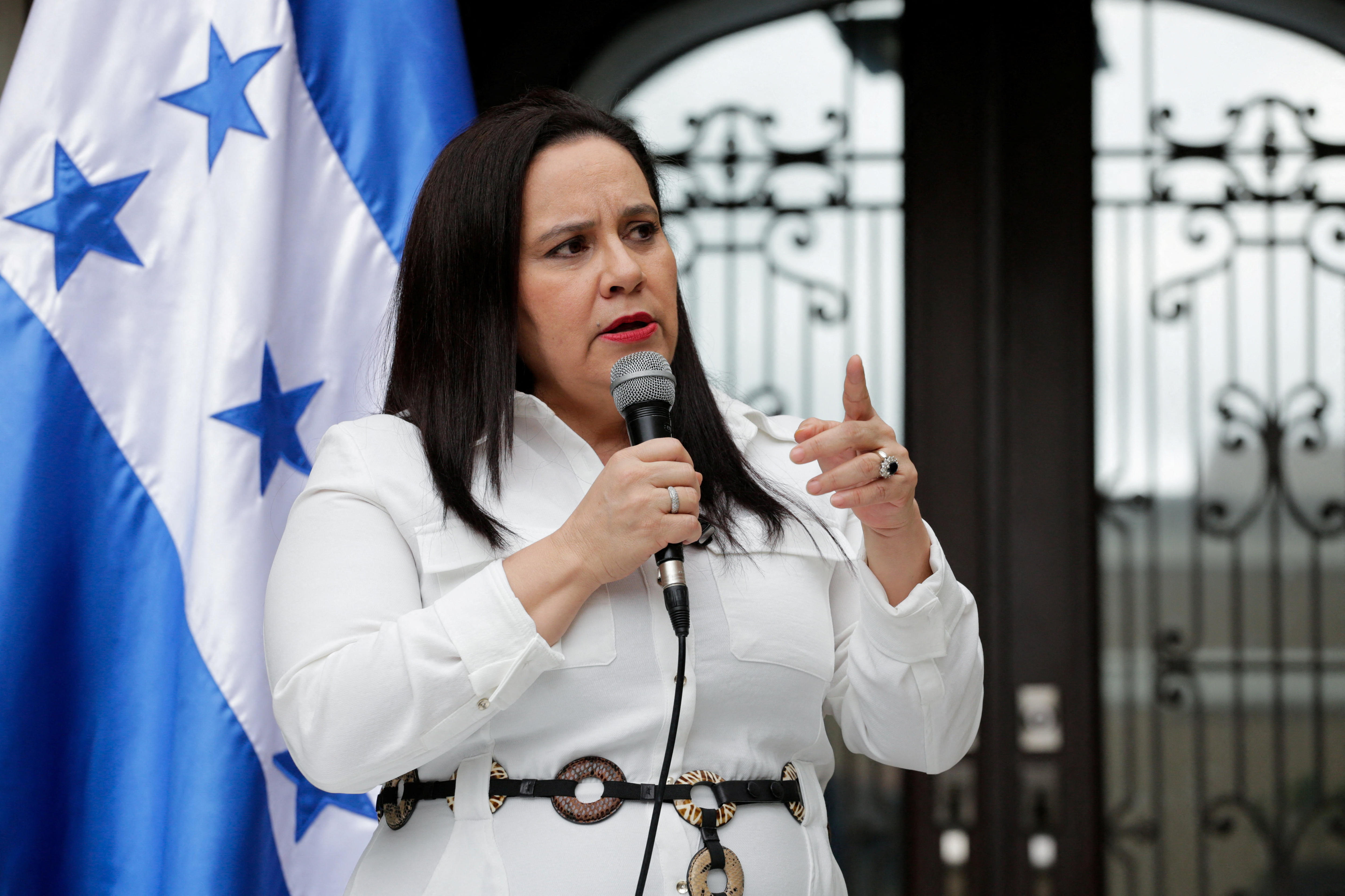 Ana Garcia holds a microphone and speaks in front of a gate and a Honduran flag