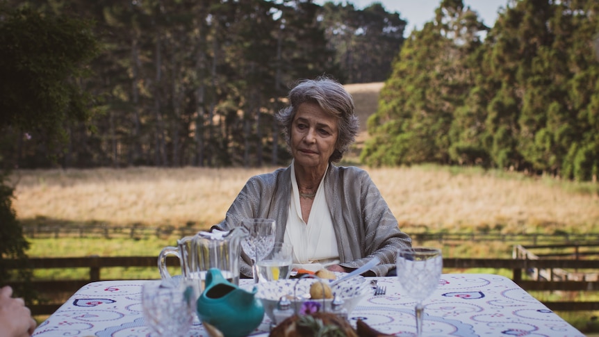 An unsmiling woman with short grey hair sits at an outdoor table set for a dinner party, with a paddock and trees behind her