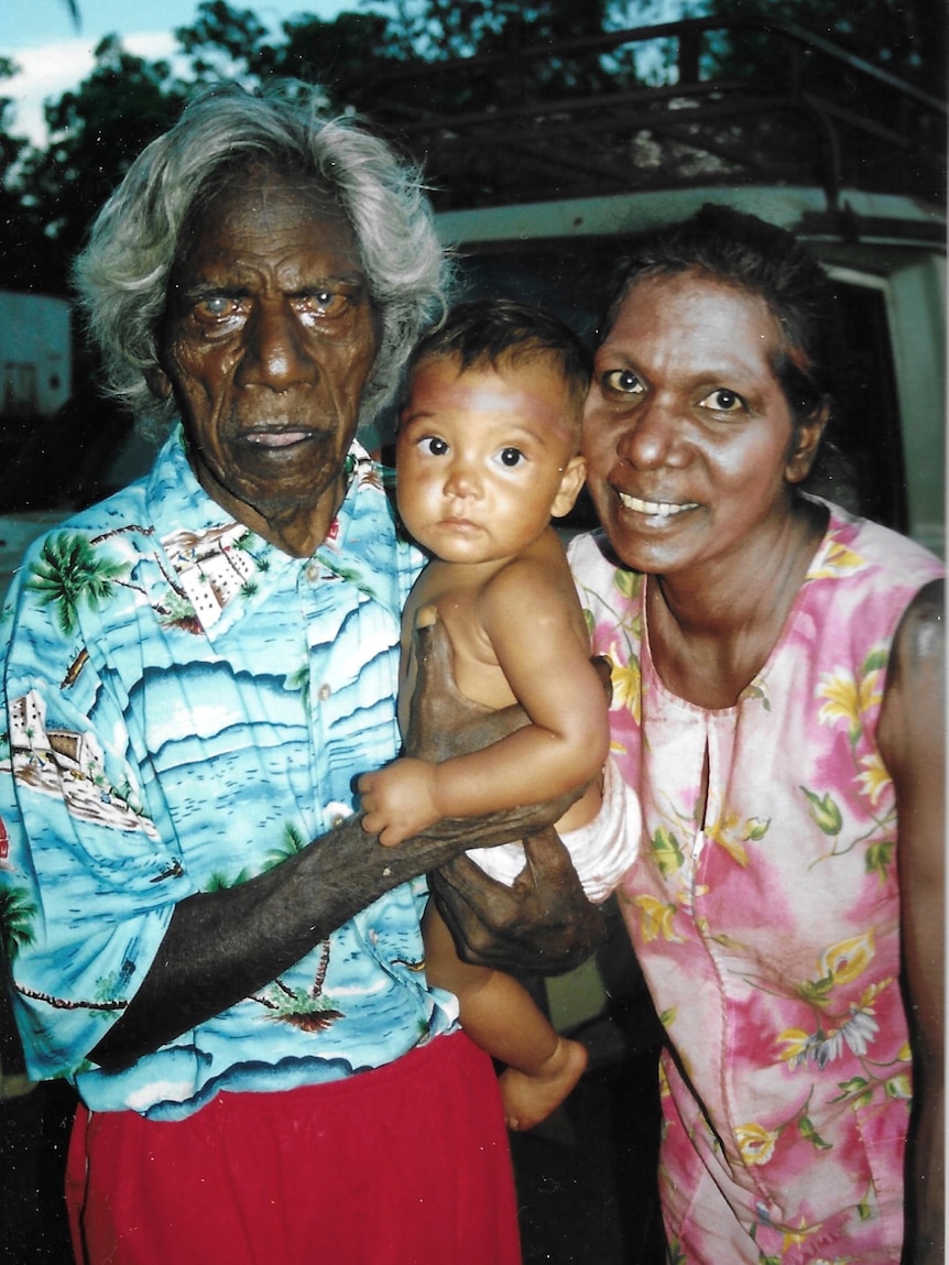 An older Indigenous man with an Indigenous woman and a Indigenous baby all look at the camera.