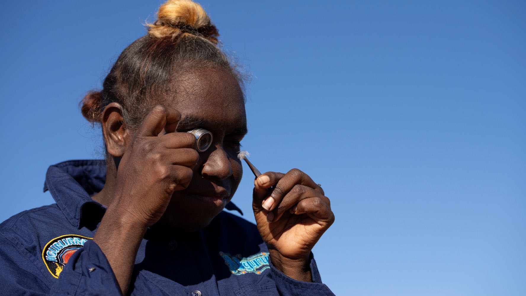 A young Aboriginal woman in dark navy long sleeve shirt holds tiny feather in tweezers and looks at it with magnifier eye piece.