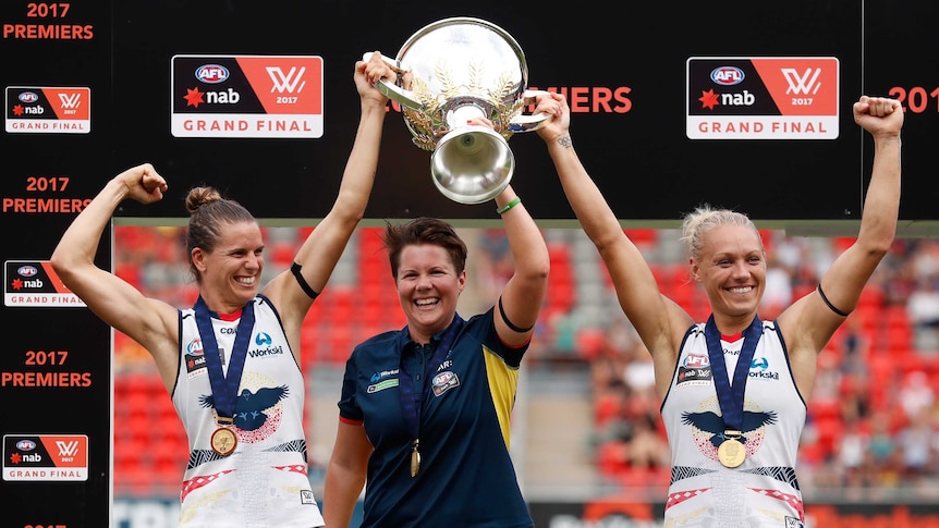 Chelsea Randall, Bec Goddard (Coach) and Erin Phillips hold up AFLW trophy.