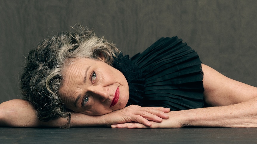 Judith Lucy, a middle-aged woman with curly, grey-streaked dark hair, rests her head on her arms, looking up.