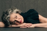 Judith Lucy, a middle-aged woman with curly, grey-streaked dark hair, rests her head on her arms, looking up.