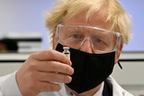 Boris Johnson holds up a vial. He is wearing goggles and a facemask.