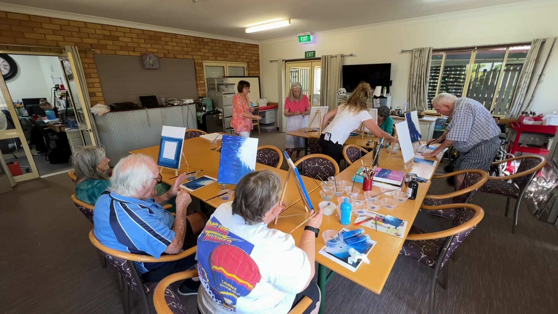 Half a dozen elderly residents sit around tables in a U shape for a painting class