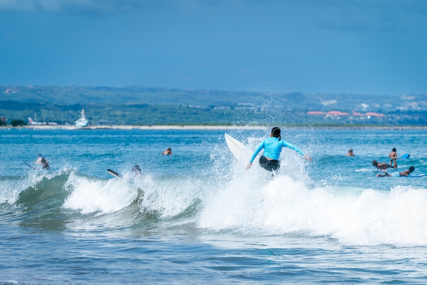A woman wearing a blue long sleeve shirt surfs a wave on her surfboard.