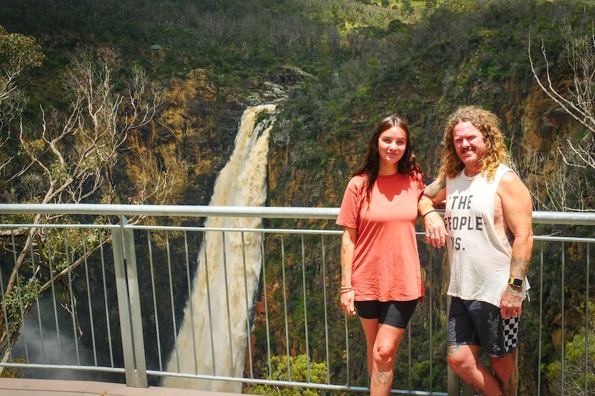 A couple pose on a platform in front of a waterfall.