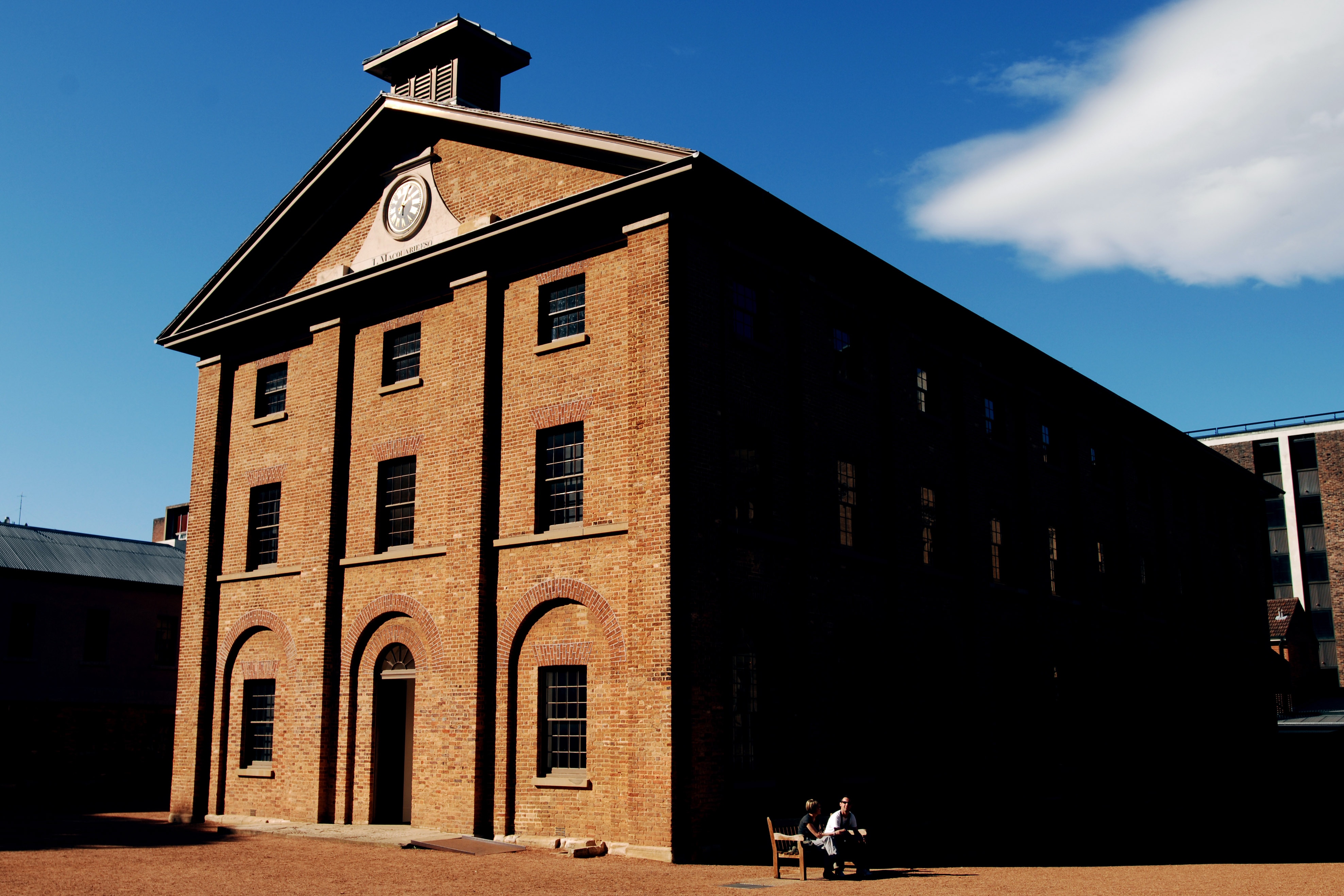A tall brown brick building with multiple stories and windows, on a sunny day. It is Hyde Park Barracks in Sydney.