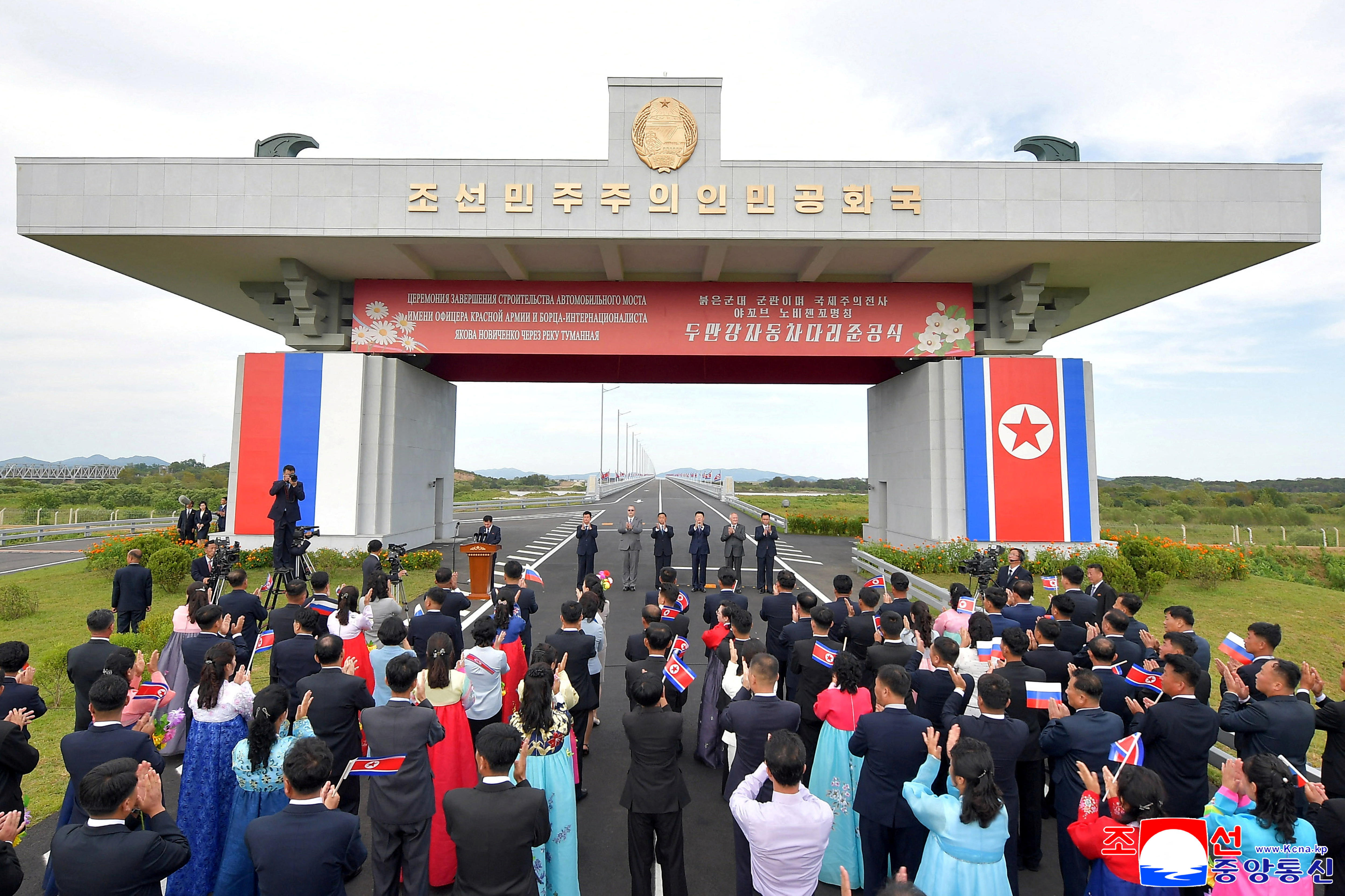 a crowd gathers around a bridge opening covered in russian and north korean flags.