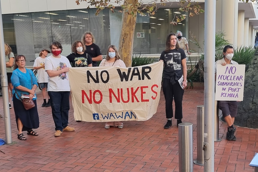 A group of people stand outside a building. Some hold a sign saying 'no war, no nukes'.