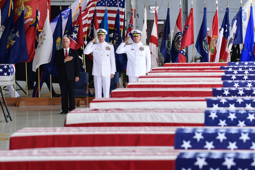 Mike Pence and military officials salute a row of cases draped with US flags