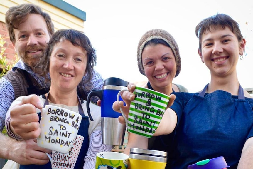 Four staff members from Saint Monday cafe hold reusable mugs to the camera.