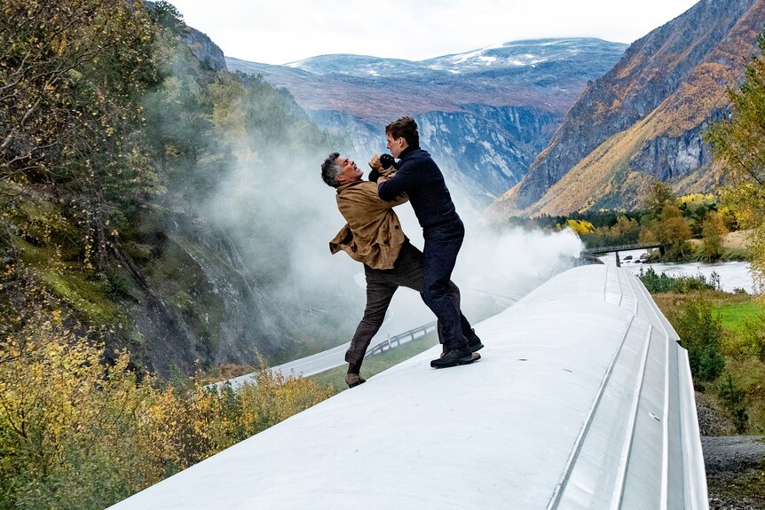 Esai Morales, a grey-haired Puerto Rican man, and Tom Cruise, a brunette white man, fight atop a train passing mountains.